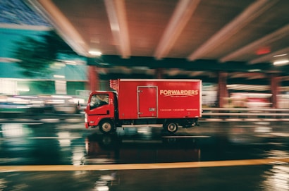 A red delivery truck is moving at high speed under an overpass on a wet road. The motion blur suggests fast movement, and the reflections on the pavement indicate recent rainfall. The truck has the word 'FORWARDERS' written on its side.