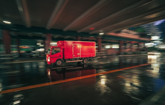 A red delivery truck moves quickly under a bridge in an urban setting at night, captured with a motion blur effect. The wet pavement reflects the lights, creating a dynamic and vibrant scene.