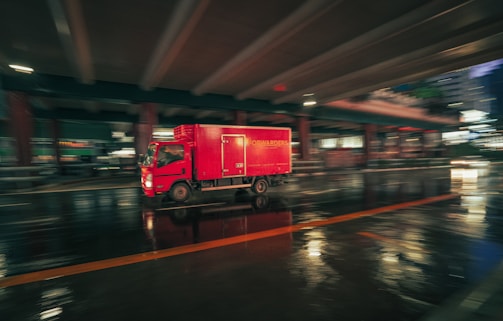 A red delivery truck moves quickly under a bridge in an urban setting at night, captured with a motion blur effect. The wet pavement reflects the lights, creating a dynamic and vibrant scene.