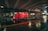 A red delivery truck moves quickly under a bridge in an urban setting at night, captured with a motion blur effect. The wet pavement reflects the lights, creating a dynamic and vibrant scene.