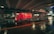 A red delivery truck moves quickly under a bridge in an urban setting at night, captured with a motion blur effect. The wet pavement reflects the lights, creating a dynamic and vibrant scene.