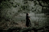 A woman stands in a lush, wooded area by a calm, reflective body of water. Her eyes are closed as she embraces herself, surrounded by intertwined branches and leaves that create a serene, natural environment. The muted green tones of the forest convey a sense of tranquility.