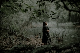 A woman stands in a lush, wooded area by a calm, reflective body of water. Her eyes are closed as she embraces herself, surrounded by intertwined branches and leaves that create a serene, natural environment. The muted green tones of the forest convey a sense of tranquility.