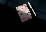 A notebook with a pink cover featuring turquoise and black insect illustrations and floral patterns is placed on a dark surface. Next to it is a pen with an intricate geometric design and a white casing.