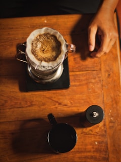 A top-down view of a coffee brewing setup on a wooden table. A freshly brewed coffee is in a glass pour-over dripper with a ceramic filter, sitting on a scale. A hand rests on the table nearby. A black cup and a coffee grinder are also on the table.