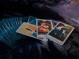 A set of tarot cards is spread out on a dark, textured surface. Three cards are prominently displayed on top of the deck, featuring vibrant and mystical illustrations. The central card showcases a figure with long red hair, surrounded by symbols and abstract designs. To the right, a card labeled 'The Hermit' depicts an antlered silhouette against a forested backdrop.