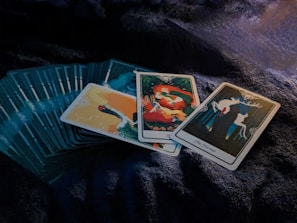 A set of tarot cards is spread out on a dark, textured surface. Three cards are prominently displayed on top of the deck, featuring vibrant and mystical illustrations. The central card showcases a figure with long red hair, surrounded by symbols and abstract designs. To the right, a card labeled 'The Hermit' depicts an antlered silhouette against a forested backdrop.