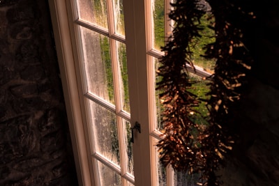 A wooden-framed glass door is adorned with green and brown garland. The door has multiple glass panels and opens to a view of lush green foliage outside. The lighting suggests that it is either sunrise or sunset, casting a warm glow on the scene.