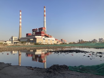 A large industrial facility is situated near the center of the image, featuring multiple structures with red and white smokestacks. A calm body of water in the foreground reflects the buildings and chimneys. The sky is clear and blue, indicating good weather. Surrounding the facility, there is barren land with some rocks and a green tarp spread across a section near the water.