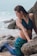 A woman dressed as a mermaid poses on a rocky beach, with wet hair and a seashell necklace. Her outfit includes a blue top and a mermaid tail, designed with blue and green scale patterns. The ocean waves crash gently on the shore behind her.
