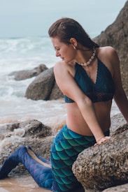 A woman dressed as a mermaid poses on a rocky beach, with wet hair and a seashell necklace. Her outfit includes a blue top and a mermaid tail, designed with blue and green scale patterns. The ocean waves crash gently on the shore behind her.