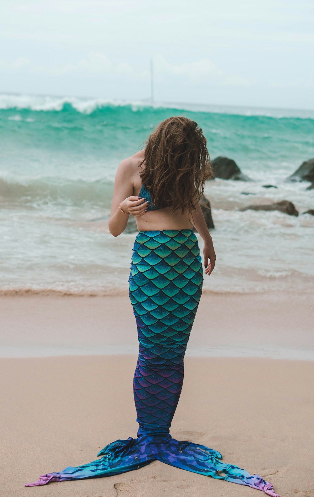 a woman in a blue mermaid tail standing on the beach