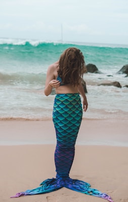 A person stands on a sandy beach wearing a colorful mermaid costume with a blue and green iridescent tail. In the background, the ocean waves gently crash against rocks.