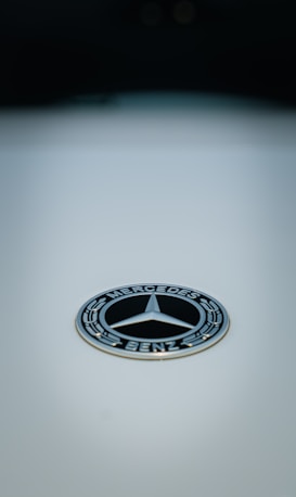 A polished silver logo featuring a three-pointed star encircled by the words Mercedes-Benz on a glossy, light background. The logo is centered, with a focus on its intricate design and metallic sheen.