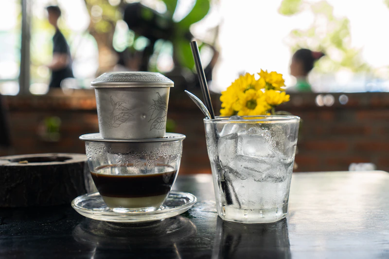 A phin filter dripping coffee beside a glass of ice water and fresh flowers
