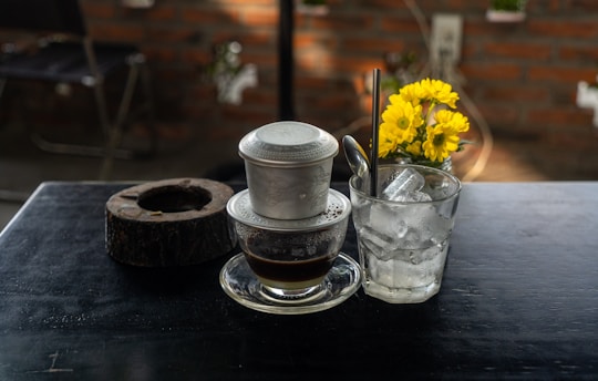 A traditional coffee filter sits atop a glass with coffee, next to a glass filled with ice and a metal spoon resting inside. Bright yellow flowers in a small vase add a splash of color against a brick wall background, and a wooden coaster lies nearby.