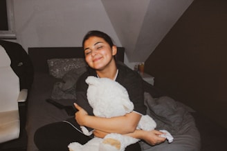 A person with closed eyes is sitting on a bed, hugging a large stuffed toy. The room has a cozy atmosphere with a white chair on the left and a nightstand on the right. A remote control and some bedding are visible.