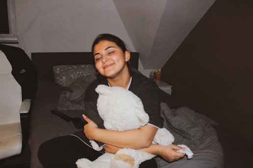 A person with closed eyes is sitting on a bed, hugging a large stuffed toy. The room has a cozy atmosphere with a white chair on the left and a nightstand on the right. A remote control and some bedding are visible.