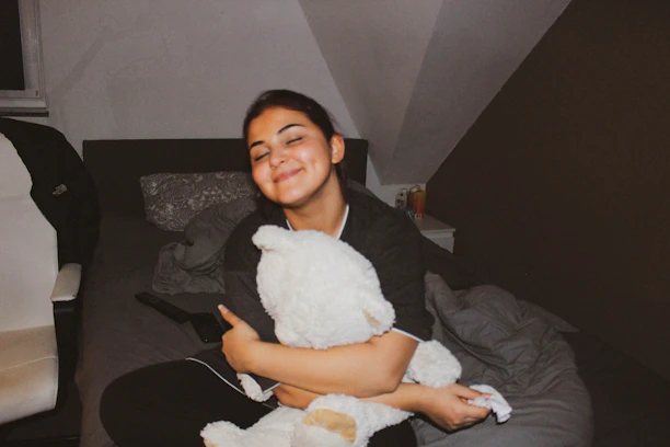 A person with closed eyes is sitting on a bed, hugging a large stuffed toy. The room has a cozy atmosphere with a white chair on the left and a nightstand on the right. A remote control and some bedding are visible.