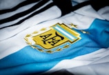 A close-up of a sports jersey featuring blue and white stripes with a golden emblem prominently displayed. The emblem includes a stylized sun and laurel leaves.