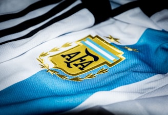 A close-up of a sports jersey featuring blue and white stripes with a golden emblem prominently displayed. The emblem includes a stylized sun and laurel leaves.
