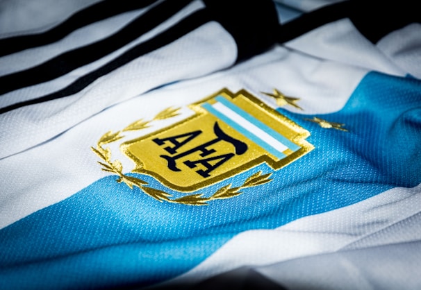 A close-up of a sports jersey featuring blue and white stripes with a golden emblem prominently displayed. The emblem includes a stylized sun and laurel leaves.