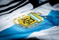 A close-up of a sports jersey featuring blue and white stripes with a golden emblem prominently displayed. The emblem includes a stylized sun and laurel leaves.