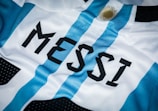 A close-up of a sports jersey featuring blue and white vertical stripes and the name 'MESSI' printed in bold black letters. A golden sun emblem is visible near the top.