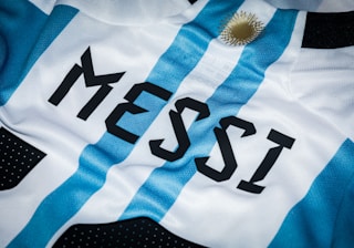 A close-up of a sports jersey featuring blue and white vertical stripes and the name 'MESSI' printed in bold black letters. A golden sun emblem is visible near the top.