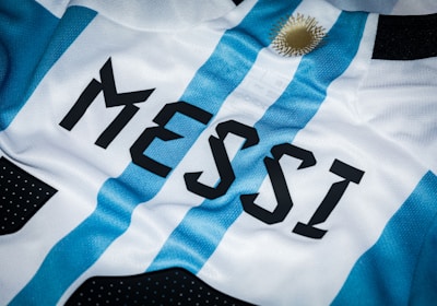 A close-up of a sports jersey featuring blue and white vertical stripes and the name 'MESSI' printed in bold black letters. A golden sun emblem is visible near the top.