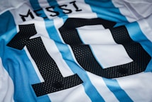A close-up of a sports jersey with blue and white vertical stripes. The number 10 is prominently displayed in black with small white dots, and the name 'Messi' is just above the number in black font.