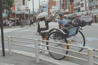A rickshaw puller wearing a traditional outfit and hat stands near a rickshaw on a city street, gesturing in conversation with a seated passenger. The urban backdrop includes storefronts, street signs, and pedestrians, creating a lively and bustling atmosphere.