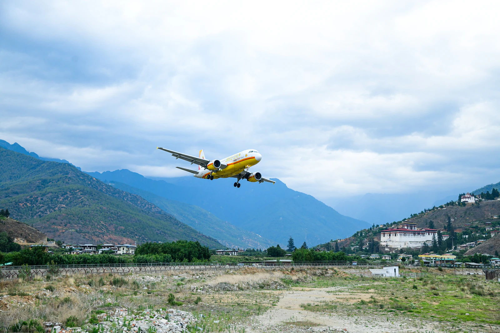 A yellow airliner flying low over Paro airport between forested ridges, Rinpung Dzong on the hill to the right