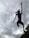 A wire sculpture of an anthropomorphic figure with animal-like features appears to leap into the air against a backdrop of a cloudy sky. The figure is highly detailed with visible whiskers and raised arms, conveying a sense of movement and dynamism.