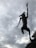 A wire sculpture of an anthropomorphic figure with animal-like features appears to leap into the air against a backdrop of a cloudy sky. The figure is highly detailed with visible whiskers and raised arms, conveying a sense of movement and dynamism.