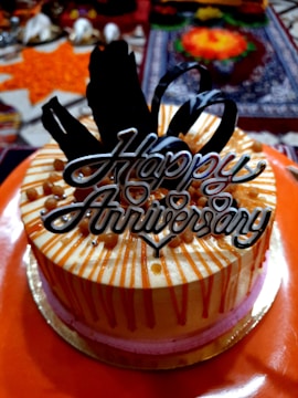 A decorated cake with orange and pink layers, topped with chocolate decorations and a Happy Anniversary sign. The background includes colorful items like flowers and a patterned fabric.