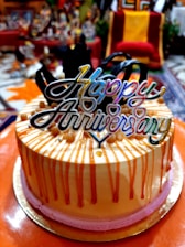 A festive cake with decorative orange drip icing and pink base trim, topped with a colorful 'Happy Anniversary' topper, surrounded by a blurred indoor setting with various objects in the background.