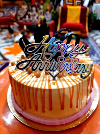 A festive cake with decorative orange drip icing and pink base trim, topped with a colorful 'Happy Anniversary' topper, surrounded by a blurred indoor setting with various objects in the background.