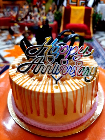 A festive cake with decorative orange drip icing and pink base trim, topped with a colorful 'Happy Anniversary' topper, surrounded by a blurred indoor setting with various objects in the background.