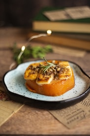 A plate with a beautifully prepared dish featuring grilled slices of cheese on top of a round piece of cooked pumpkin, garnished with herbs and spices. The setting includes a rustic wooden table, some old papers, and blurred books in the background, with subtle fairy lights adding a cozy ambiance.