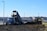 A blue dump truck is tipping its load of dirt onto a construction site, with a yellow bulldozer nearby preparing to spread the material. The setting is an open area with commercial buildings and a clear blue sky in the background.