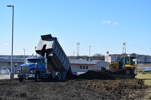 A blue dump truck is tipping its load of dirt onto a construction site, with a yellow bulldozer nearby preparing to spread the material. The setting is an open area with commercial buildings and a clear blue sky in the background.