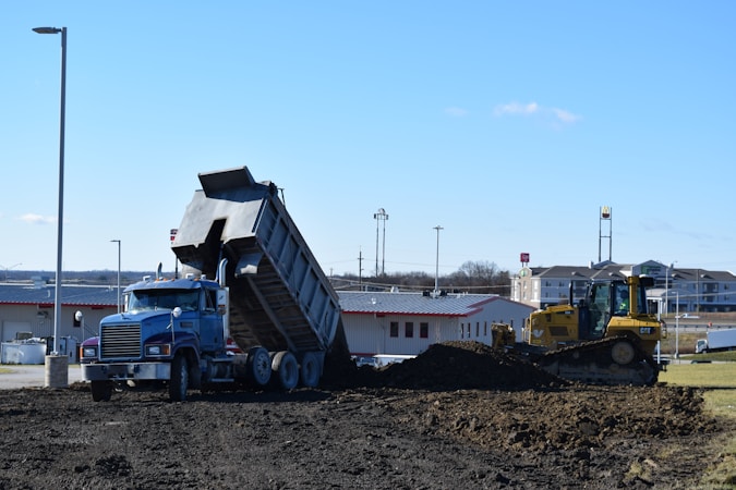 A blue dump truck is tipping its load of dirt onto a construction site, with a yellow bulldozer nearby preparing to spread the material. The setting is an open area with commercial buildings and a clear blue sky in the background.