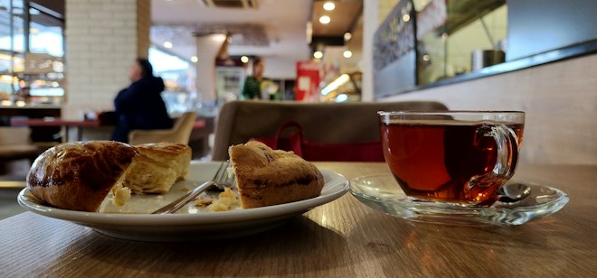 A cozy indoor caf&eacute; setting with a focus on a wooden table where a plate with a sliced pastry and a clear glass cup of tea are placed. The background features a blurred view of people sitting, with warm lighting and decorative elements.