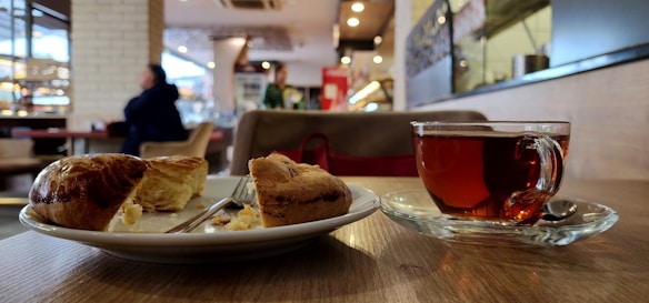 A cozy indoor café setting with a focus on a wooden table where a plate with a sliced pastry and a clear glass cup of tea are placed. The background features a blurred view of people sitting, with warm lighting and decorative elements.