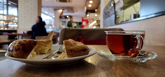 A cozy indoor caf&eacute; setting with a focus on a wooden table where a plate with a sliced pastry and a clear glass cup of tea are placed. The background features a blurred view of people sitting, with warm lighting and decorative elements.