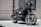 A sleek black motorcycle with a quilted seat and a large front headlight is parked in front of a building with beige brick walls and a curtained window. The motorcycle has distinctive branding, polished chrome elements, and an overall modern, custom design.