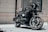 A sleek black motorcycle with a quilted seat and a large front headlight is parked in front of a building with beige brick walls and a curtained window. The motorcycle has distinctive branding, polished chrome elements, and an overall modern, custom design.