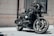 A sleek black motorcycle with a quilted seat and a large front headlight is parked in front of a building with beige brick walls and a curtained window. The motorcycle has distinctive branding, polished chrome elements, and an overall modern, custom design.