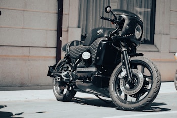 A sleek black motorcycle with a quilted seat and a large front headlight is parked in front of a building with beige brick walls and a curtained window. The motorcycle has distinctive branding, polished chrome elements, and an overall modern, custom design.